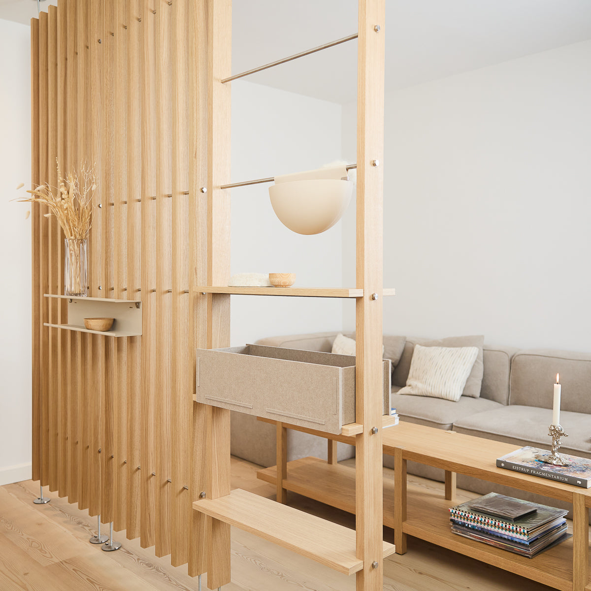 Modern living room with a Nordic light wood slatted EMBRACE room divider, beige sofa, coffee table, and accents including books, a candle, and dried flowers.